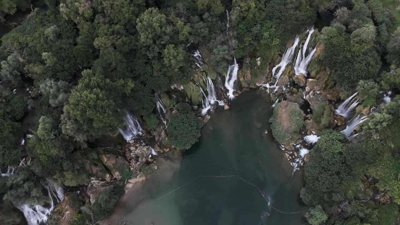 Cascate a più livelli che si gettano in una piscina naturale verde incorniciata da una fitta foresta, ripresa con drone