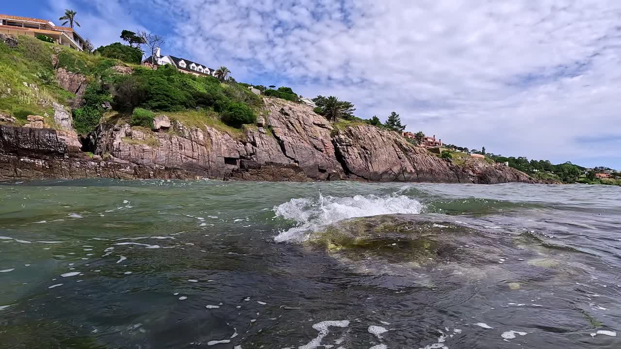 olas salpicando contra la cámara, mostrando mansiones en la cima de la colina, en punta ballena, uruguay