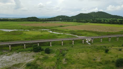 angled aerial sliding footage towards the left revealing the elevated railways, farmland with cattle, mountains and lake in the horizon, muak klek, saraburi, thailand