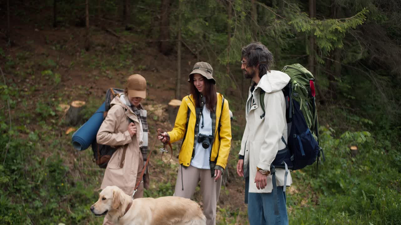un trío de excursionistas en ropa especial de senderismo con grandes mochilas, junto con su perro de color claro, se comunican en el medio del bosque