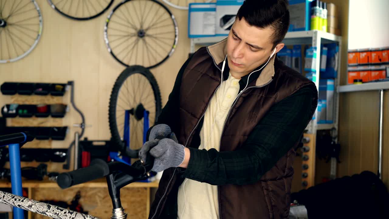 un empleado ocupado está desmontando el manillar de la bicicleta y limpiando las piezas con un paño mientras repara la bicicleta en el taller. un joven está escuchando música con auriculares.