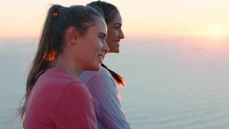 Amigas-Sentadas-En-La-Cima-De-La-Montaña-Mirando-La-Tranquila-Vista-Del-Océano-Al-Atardecer-Dos-Mujeres-Descansando-Después-De-Una-Caminata-Disfrutando-De-Una-Tranquila-Aventura-De-Viaje-Al-Aire-Libre