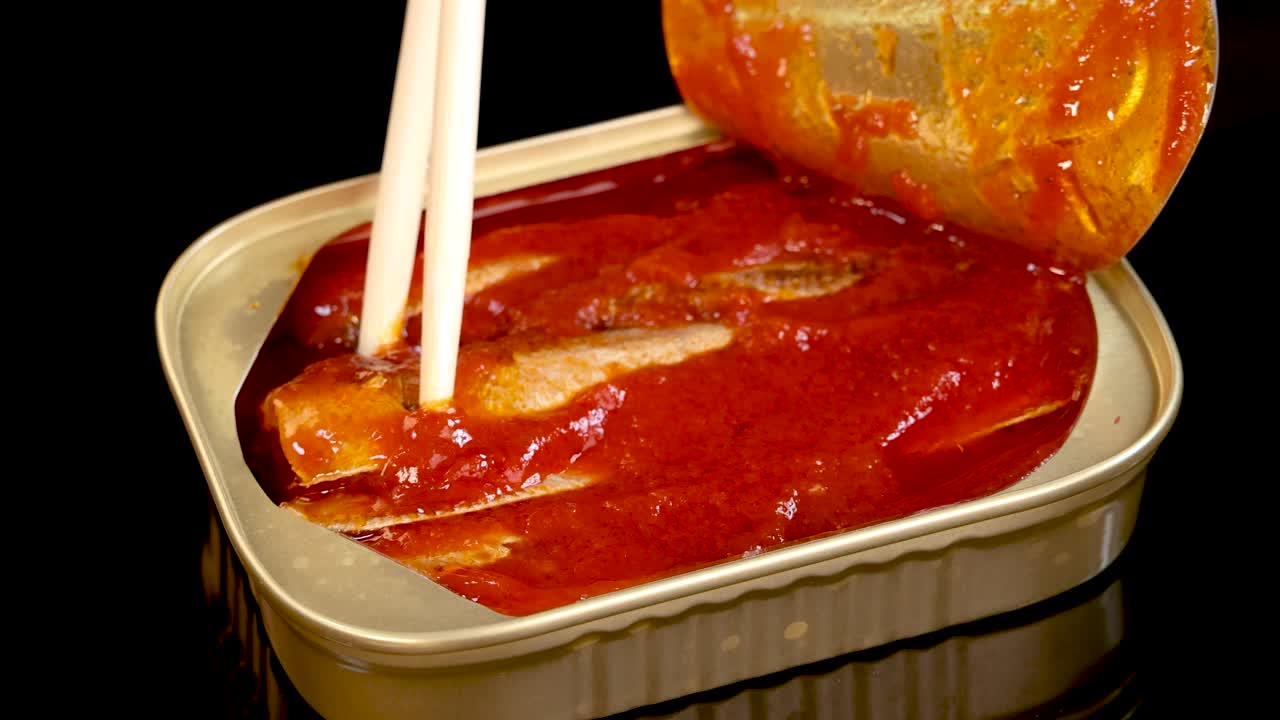 Chopsticks grasp and lift a sardine from an open can filled with tomato sauce, under bright, direct lighting with a black background and minimal camera movement