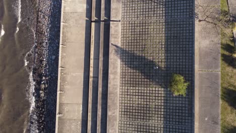 toma de seguimiento aérea hacia abajo de una persona que se despierta en un suelo pedregoso junto a la orilla del río con olas que alcanzan al atardecer