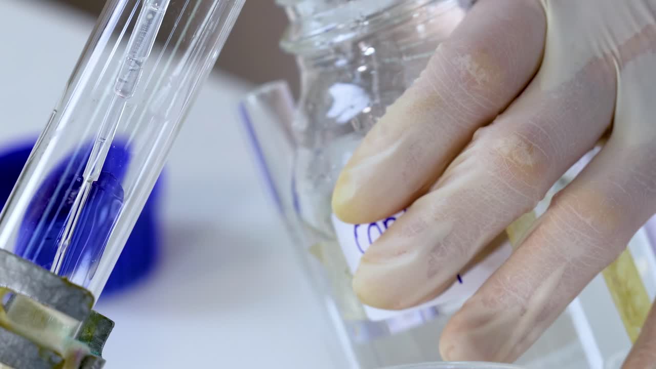Close-up of gloved hands manipulating glassware with liquid in a laboratory setting.