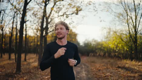 A-happy-confident-man-with-curly-hair-and-a-beard-in-a-black-sports-uniform-with-a-watch-on-his-wrist-runs-during-his-morning-jog-in-a-sunny-morning-autumn-forest-along-the-earth-path