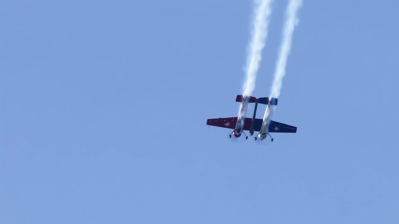 A twin-engine aircraft performs vertical maneuvers with smoke trails against a clear blue sky.