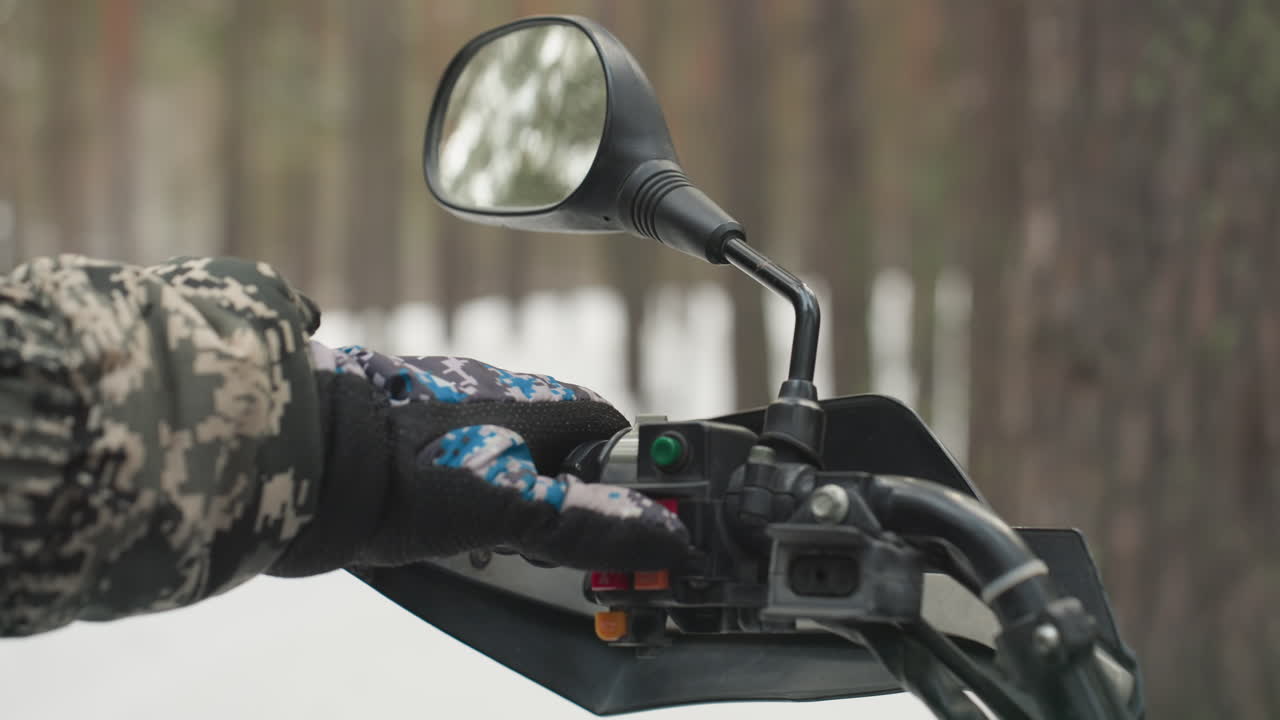 Mão enluvada no guidão na neve, jaqueta camuflada testando acelerador e interruptores, espelho refletindo floresta de pinheiros, close-up da pegada e ignição, preparação focada para passeio off-road extremo.