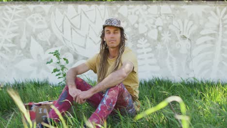 video portrait of smiling caucasian man with dreadlocks sitting on grass by mural on wall