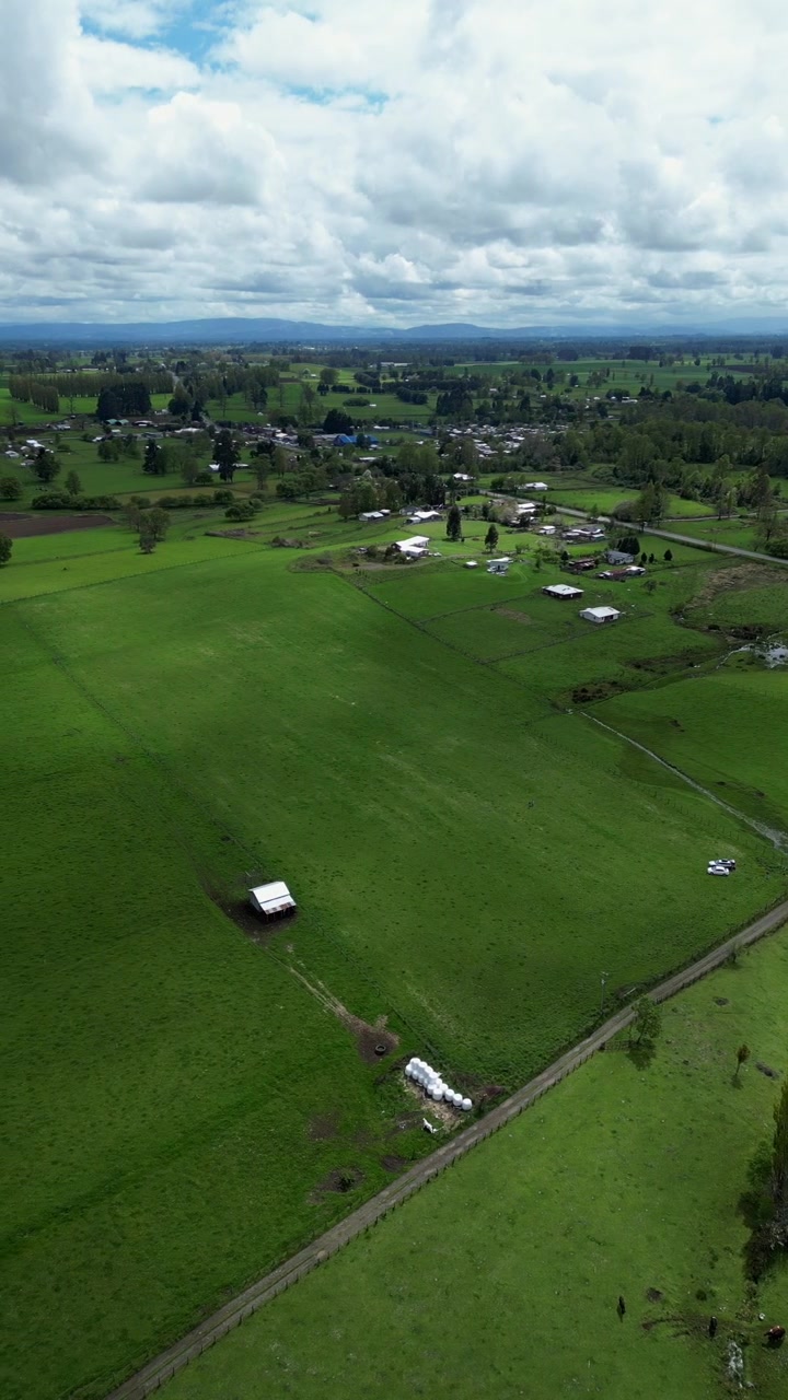 Panoramic pan over vast green farmland with scattered rural homesteads fields and a small town stretching toward distant mountain ranges under dramatic cloudy skies