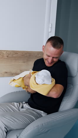 happy smiling man sits in a chair waving a newborn. loving and caring dad canoodling his baby. vertical video