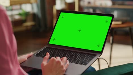 mujer adulta joven trabajando desde el apartamento del loft de la casa en una computadora portátil con pantalla verde. mujer creativa usando trackpad y navegando por internet. close up sobre el hombro.