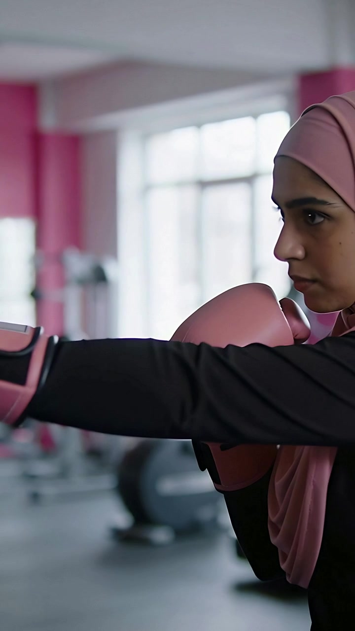Vertical video: Starting drill, woman throwing jabs at gym wearing pink hijab and gloves