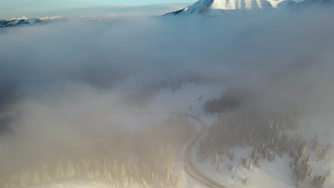 Fliegen-über-Wolken-Und-Malerische-Route-Am-Teton-Pass-An-Sonnigen-Winterabenden,-Schneebedeckte-Gipfel-Und-Hügel,-Drohnenaufnahme