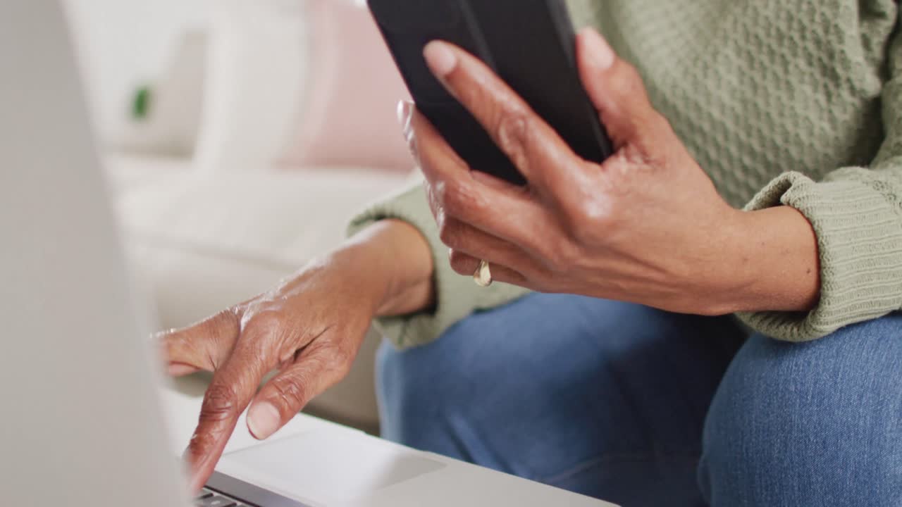 retrato de una mujer afroamericana mayor sentada en el sofá, usando una computadora portátil y un teléfono inteligente