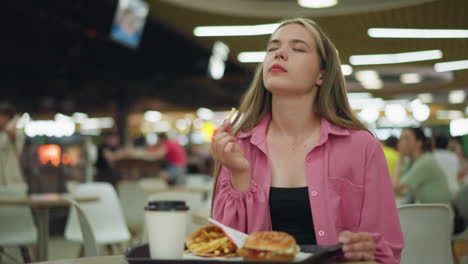 woman sits in a restaurant enjoying her meal of fries, burger, and coffee, she takes a bite of the potato chips, closes her eyes in satisfaction, and shakes her body happily, savoring the moment