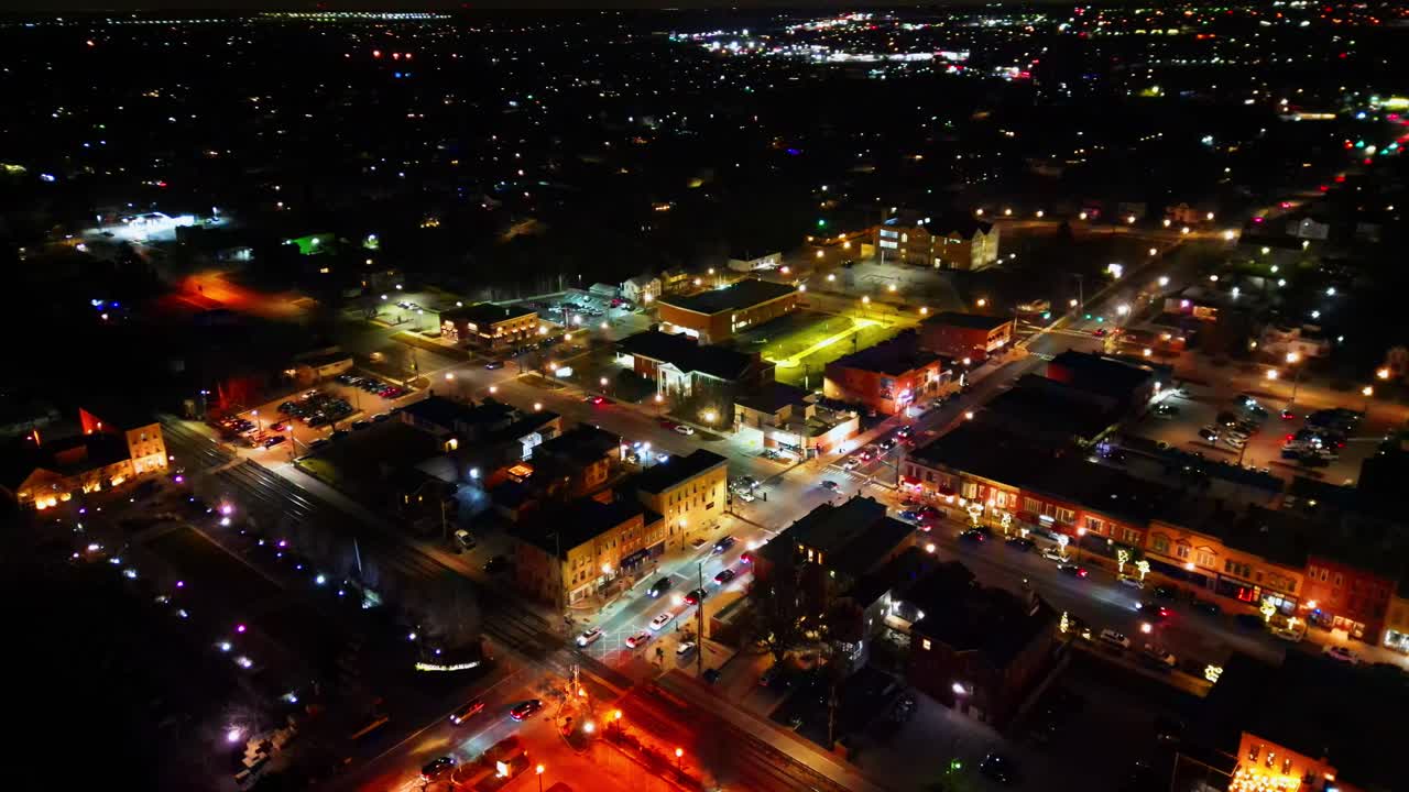 An aerial drone perspective captures the vibrant downtown of Lockport, Illinois, at night, with streetlights, car headlights, and glowing businesses illuminating the active urban landscape