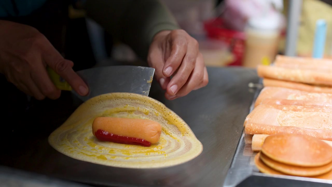 Close-up de mãos enrolando uma panqueca de salsicha com ketchup numa chapa de metal plana