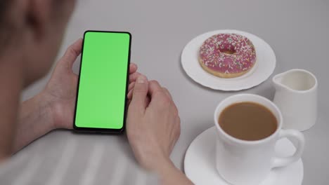 woman sitting at table coffee donut using smartphone with chroma key green screen, scrolling through social network media online shop internet. smartphone in horizontal mode with green screen mock-up.