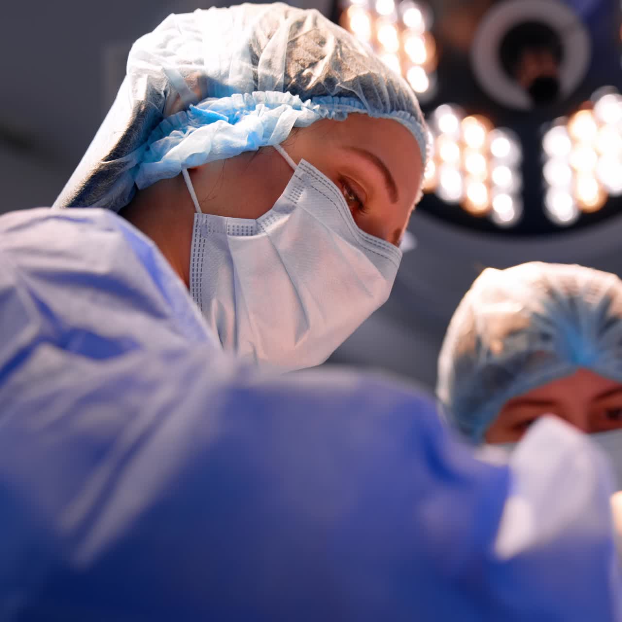 Chirurgo donna sicura di sé in uniforme blu, cuffia e mascherina che opera il paziente. Inquadratura dal basso sui medici concentrati sul lavoro in sala operatoria