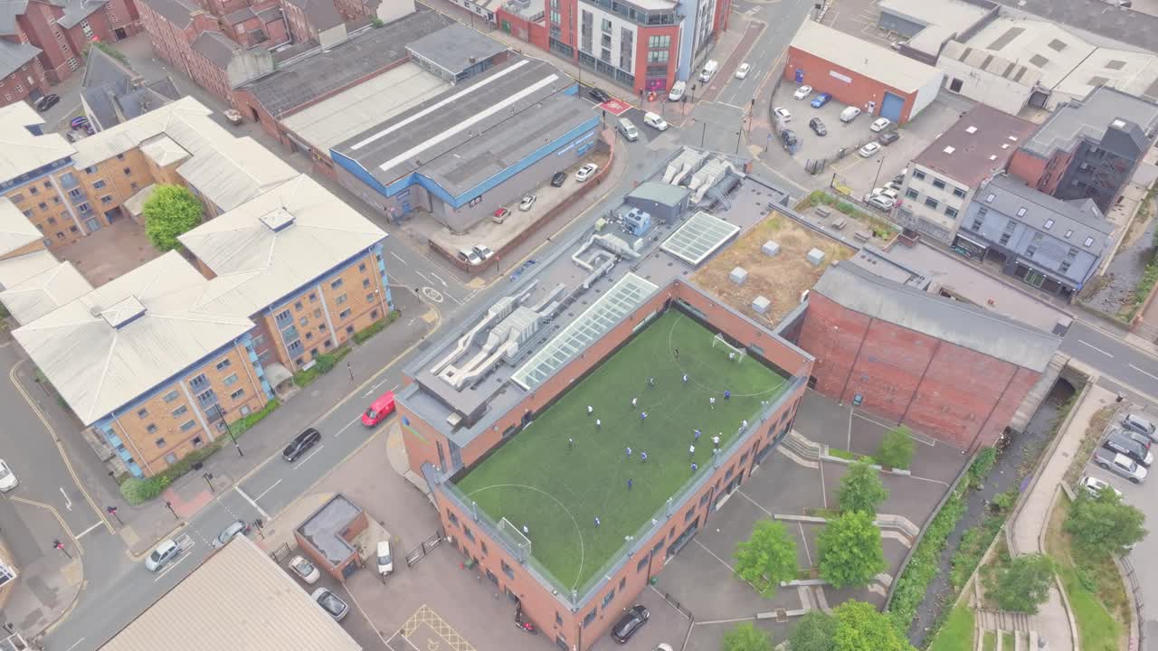 Behold the vibrant cityscape from above, featuring a mix of residential and commercial buildings, bustling streets lined with cars, and a refreshing touch of green from a soccer field on the rooftop