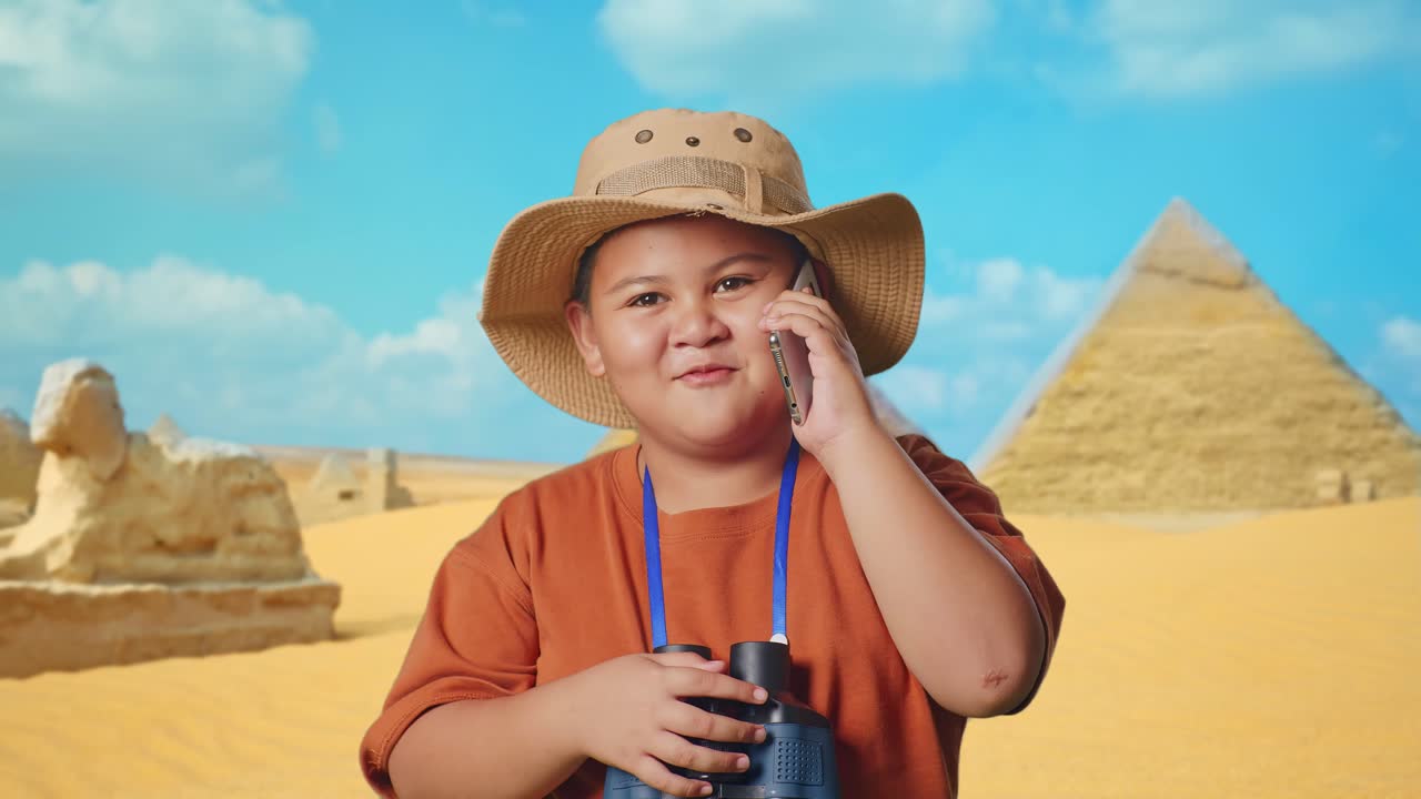 Asian Boy With A Hat And Binoculars Talking On Smartphone While Traveling In Giza Pyramid. Boy Researcher Examines Something, Travel Tourism Adventure Concept, Close Up