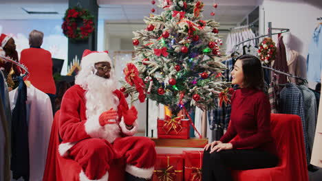 dolly out shot of worker acting as santa claus sitting down next to christmas tree with asian woman in xmas decorated clothing store. fashion shop retail assistant offering present to customer