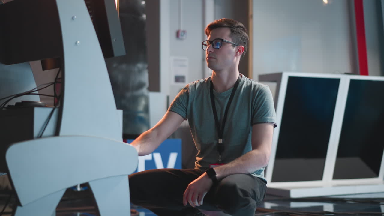 Man working in a studio, adjusting video production equipment, kneeling beside a monitor. Studio setup with bright lighting, reflective floors, and technical equipment in the background