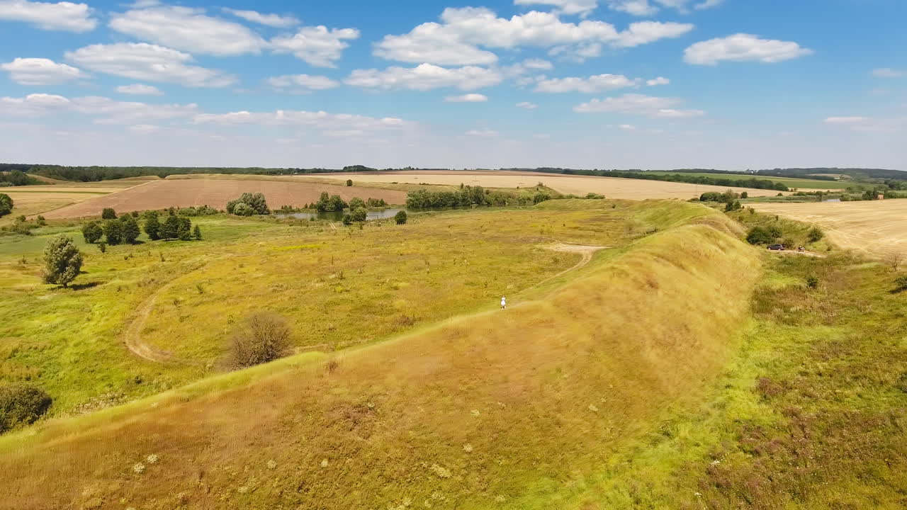 Sunny scenery of natural fields and agricultural plantations. Long hill in the meadow with thin path on top. Woman walking along the path.