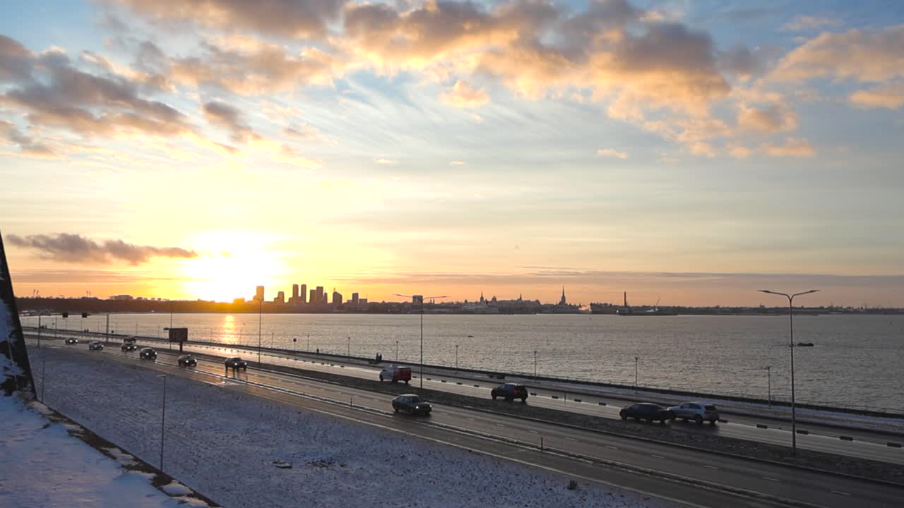 타린 시의 실루과 피리타 도로에서 자동차와 차량이 운전하는 동안 오렌지 황금빛의 일몰이나 일출이 도시의 높은 건물 뒤에서 일어나고 있습니다. 발트 반사 해는 오른쪽에 볼 수 있습니다.