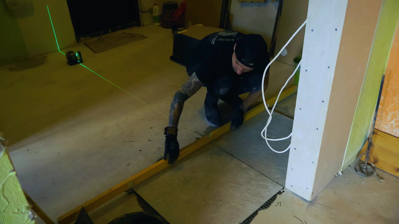 Skilled construction worker spreading adhesive with a notched trowel and carefully placing large ceramic floor tiles during a home renovation project, using a laser level for precision