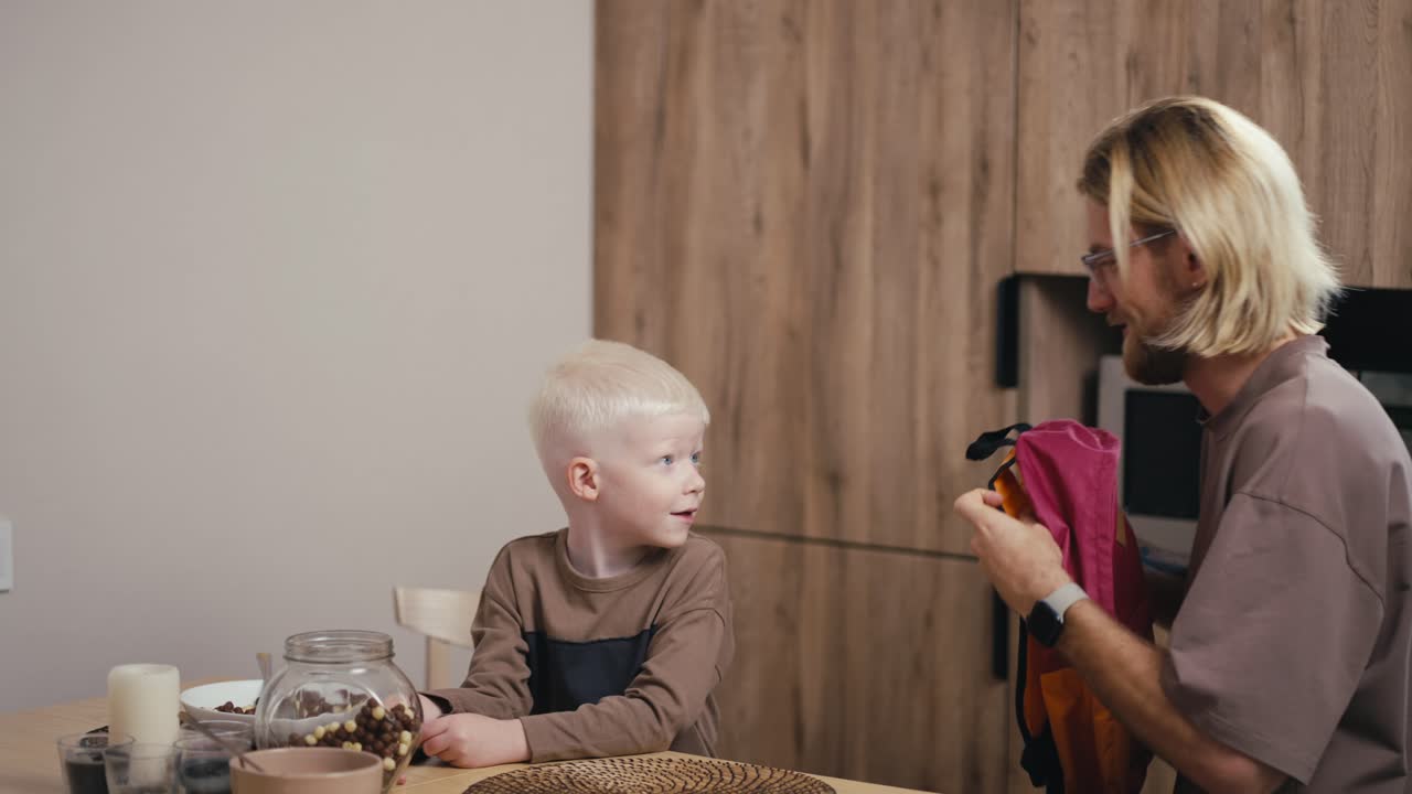 un niño albino feliz con cabello blanco se pone una mochila brillante antes de ir a la escuela en la cocina y su padre rubio con barba y gafas le ayuda con esto