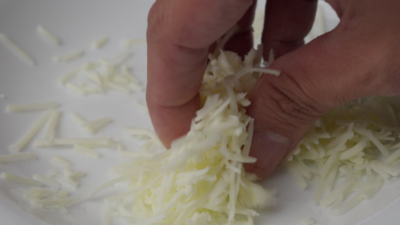 Hand reaches for a small pile of shredded cheese on a white plate for cooking or topping.