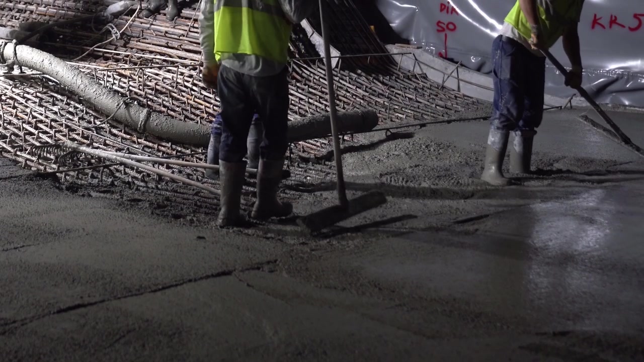 Construction workers in safety boots leveling fresh concrete over steel rebar grid during a night shift [cite: AA_17.jpg]. Detail shot of industrial slab pouring and site operations
