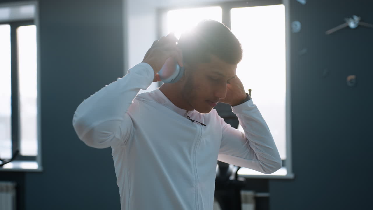 Sport man in white workout wear removes headphones with confident expression inside gym filled with natural sunlight, bright glow around head, modern equipment and large windows visible in background