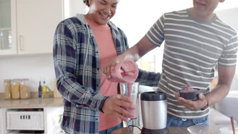 una pareja gay feliz y diversa vertiendo un batido de frutas saludable en la cocina, en cámara lenta.