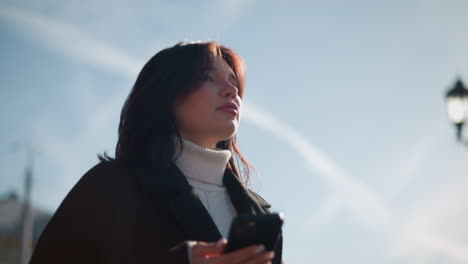 lady in black coat and white turtleneck outdoors holding smartphone, sunlight reflecting on her with blurred background featuring a lamp pole