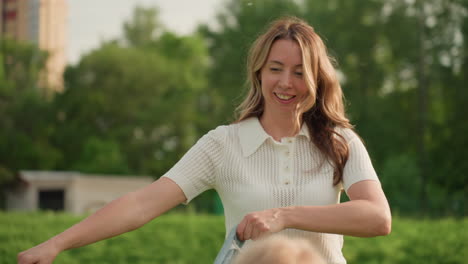 beautiful mother holding picnic blanket while smiling playfully in sunny park, casual white top, warm light on hair, background green grass, joyful relaxed moment before family picnic