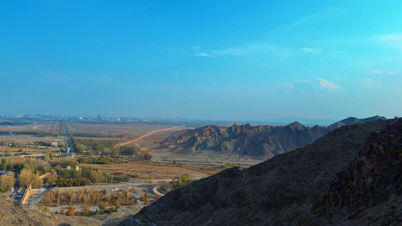 La Grande Muraglia a strapiombo, città di Jiayuguan, provincia di Gansu, Cina - Ampia veduta di aspre colline e una valle aperta che si estende verso una città distante sotto un vasto cielo blu - Panoramica a destra