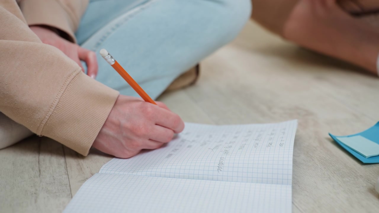 Hvid kvinde skriver grammatiknoter indendørs, sidder med krydsede ben på gulvet i hyggelig sweater og denim, blyant markerer notesbogssider, sticky notes og etui i nærheden, stille fokuseret tutor-øvelse