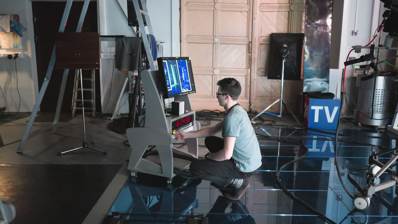 Man adjusting settings on digital equipment and monitor in modern studio during production, crouching near high-tech setup with timer displayed on screen, professional filming environment