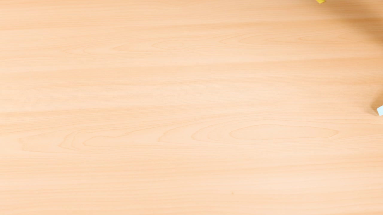 Person clears blue origami paper scraps from light wooden table, overhead view, even lighting