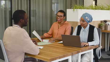 an woman and a muslim woman co workers interview a young man sitting at a table in the office 4