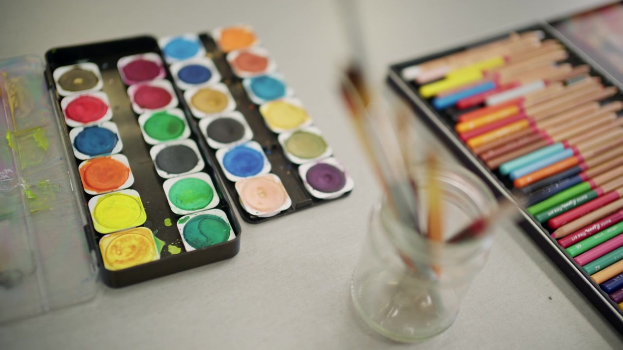 Close up of paintbrushes in a glass jar with watercolor palette and colored pencils blurred in the background