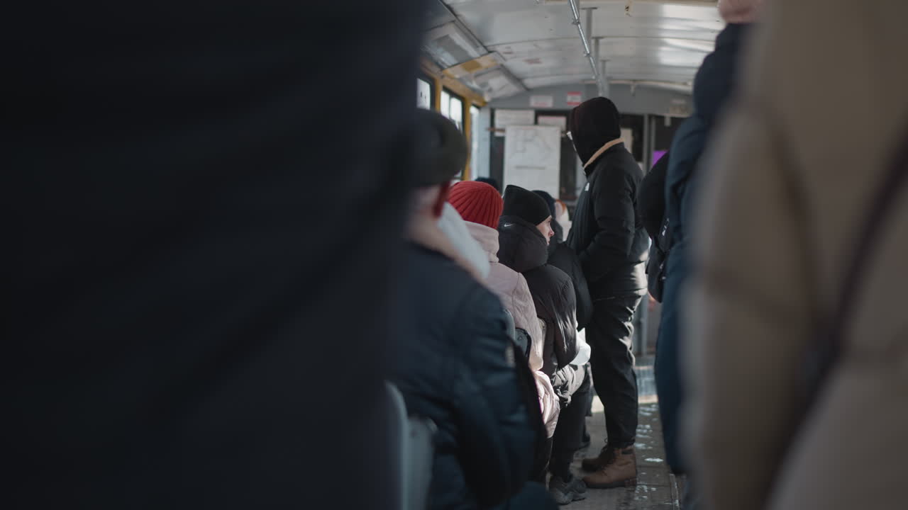 Back view of bundled passengers seated inside moving vehicle during winter commute, coats and hats visible, rider standing near aisle, cold light through windows, quiet urban ride with rows of seats