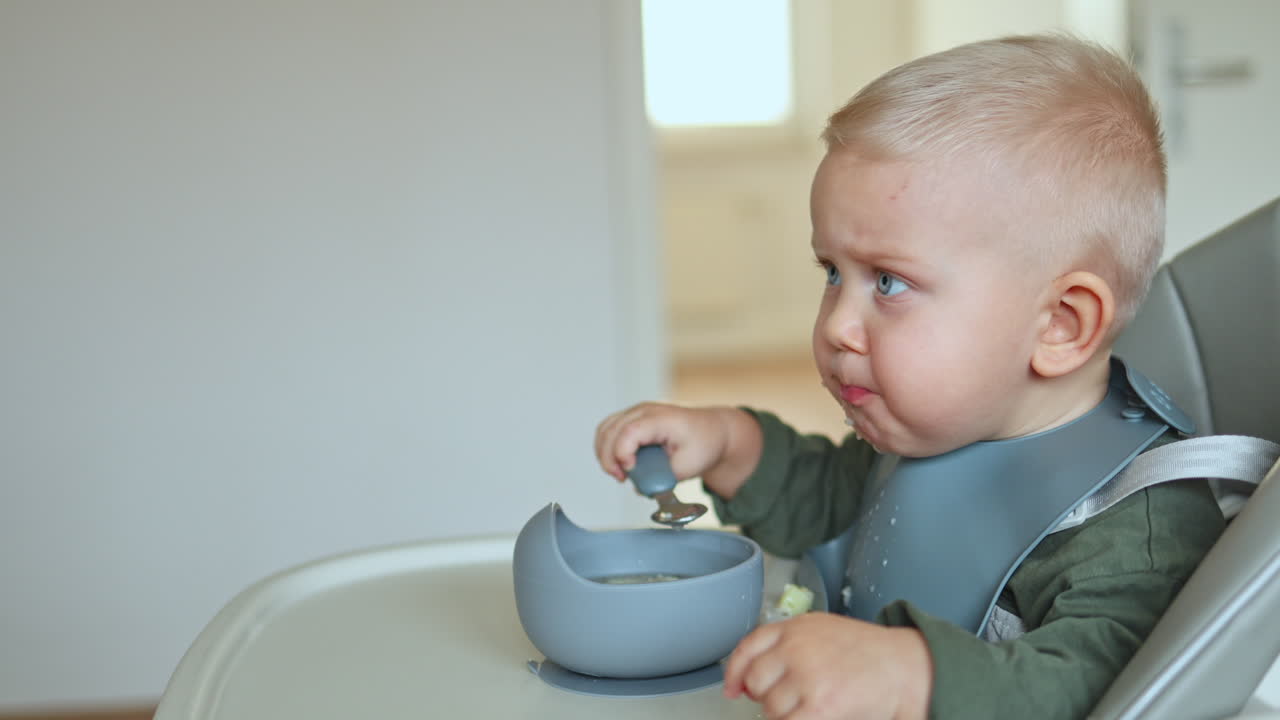 One-year-old boy sits in a high chair and learns to eat by himself. Silicone tableware for children. Boy eats from a plate by himself