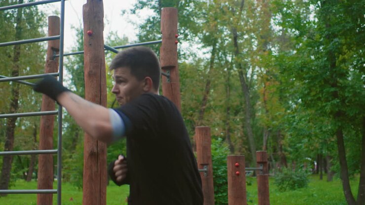 white man boxing outdoors in park performing focused shadowboxing and punch drills near wooden calisthenics bars, wearing black gloves and athletic gear, calm overcast sky, green trees, gritty