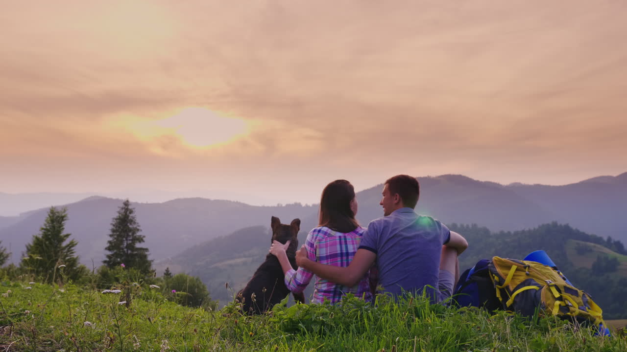개를 데리고 있는 관광객 두 명이 땅에 앉아 산의 아름다운 경치를 감상한다