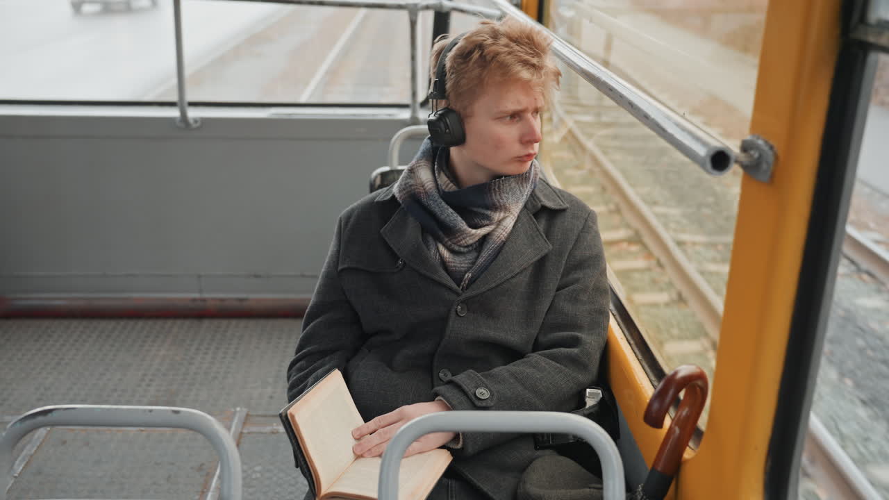 Ung hvit skaper skisserer på trikken. Notatbok åpen på fanget, oppmerksomt blikk mot vinduet, hodetelefoner på, byrute synlig gjennom glasset, penn i hånden, en observerende kunstnerstemning, reisestudiescene
