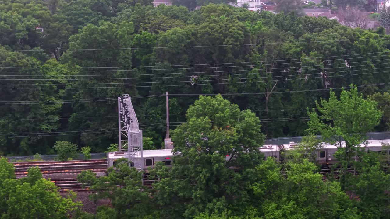An aerial view of a Long Island Rail Road train traveling on a cloudy day. The drone camera tilted downward, dolly out and truck left then pan left to follow the train moving through green trees.